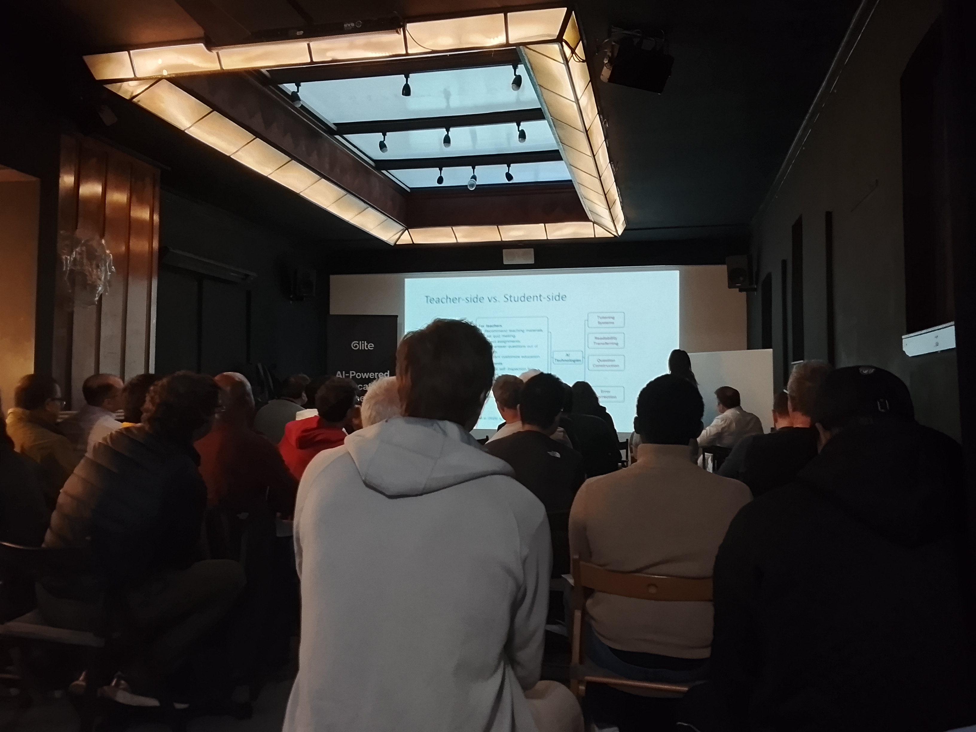 An audience at Anomalous Space watches an April meetup presentation comparing teacher-side and student-side uses of AI.